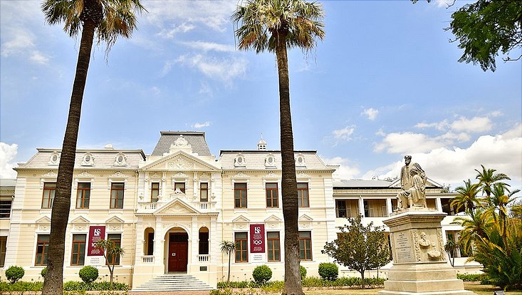 weißes Hauptgebäude der Universität Stellenbosch hinter Palmen und vor blauem Himmel