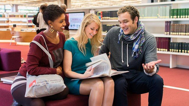 Drei junge Menschen unterschiedlicher Hautfarbe sitzen in einer Bibliothek und unterhalten sich.