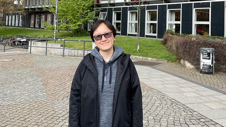 Person in Grauem Pullover, schwarzer Jacke und Jeans vor einem modernen Gebäude mit vielen Fenstern