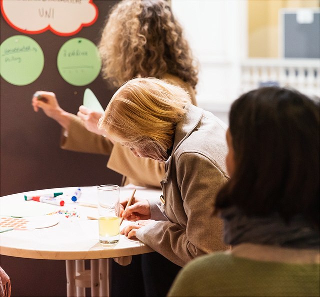 Ein Foto zeigt eine Gruppe von Menschen, die an einem runden Tisch stehen und arbeiten. Eine ältere Frau mit blonden Haaren ist im Vordergrund zu sehen, wie sie sich konzentriert und etwas auf ein Blatt Papier schreibt.