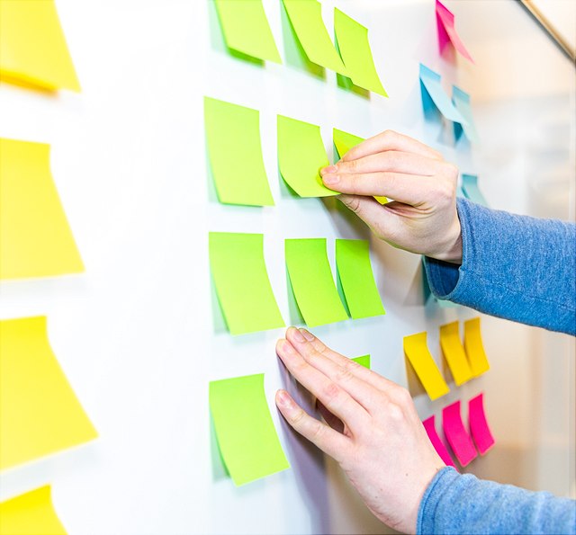Ein Foto zeigt eine Person, die bunte Haftnotizen an eine weiße Tafel anbringt. Die Person trägt ein blaues Langarmshirt. Es sind Haftnotizen in verschiedenen Farben wie Grün, Gelb, Blau und Pink zu sehen, die in geordneten Reihen angeordnet sind.
