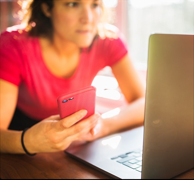 Das Foto zeigt eine Frau in einem roten T-Shirt, die an einem Laptop arbeitet. Sie hält ein rotes Smartphone in der Hand und schaut konzentriert auf den Laptop-Bildschirm.