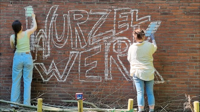 Zwei Personen schreiben das Wort Wurzelwerk an eine Klinkerwand