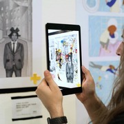 Frau schaut durch iPad auf ein Bild. Auf dem iPad ist das Bild als Augmented Reality mit Zusatzinformationen angezeigt.