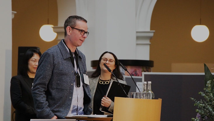 4a-733x414 Mann am Sprechpult. Zwei Frauen im Hintergrund schauen ihn an.