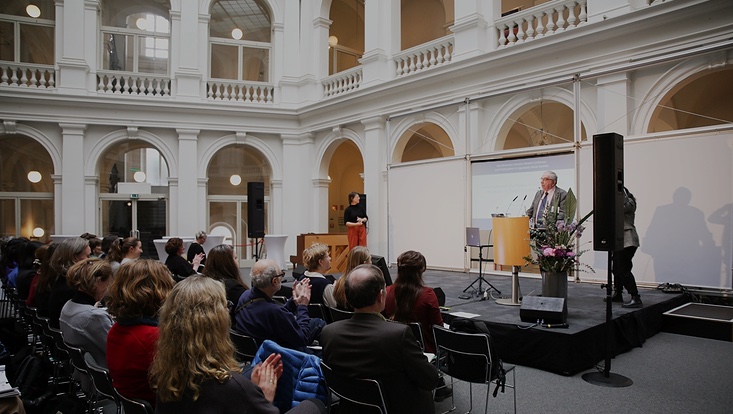 13-733x414 Person spricht auf Bühne, Publikum klatscht. Blick von hinter dem Publikum.