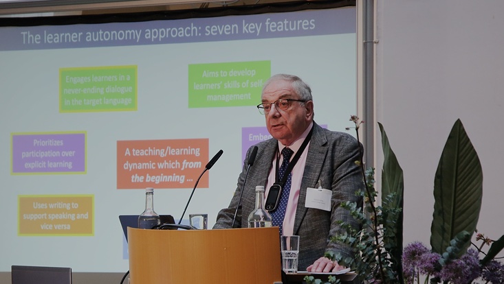 14b-733x414 Person spricht auf einer Bühne in ein Mikrofon. Präsentation im Hintergrund.