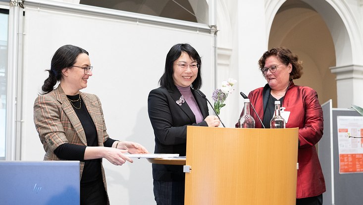 29b-733x414 Drei Frauen auf einer Bühne vor einem Sprecherpult.