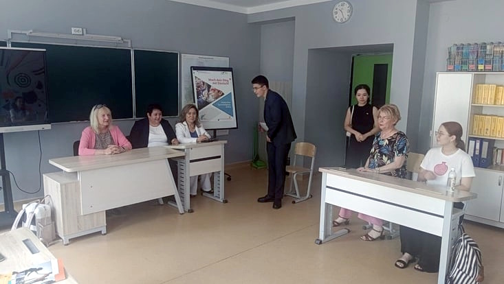 Telse Iwers, Dilbar Ernazarova und eine usbekische Kollegin sitzen an einem Lehrerpult vor einer Tafel und blicken auf einen Klassenraum mit anderen Personen. sie werden von einem Mann begrüßt