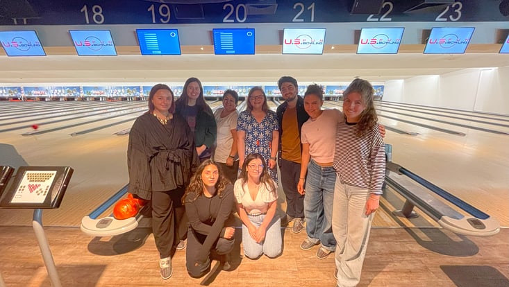 Teilnehmende stehen als Gruppe zusammen und lächeln in die Kamera. Hinter Ihnen sind mehrere Bahnen eines Bowlingcenters zu sehen.