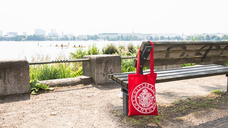Blick auf die Alster mit Sitzbank, an der ein roter Beutel mit dem Logo der Uni Hamburg hängt - dekorativ