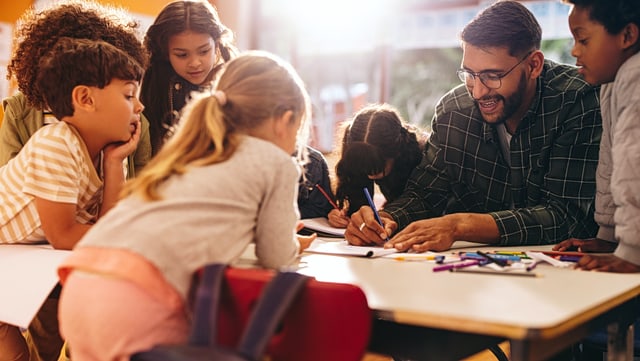Lehrer schreibt etwas umgeben von Kindern unterschiedlicher Ethnien