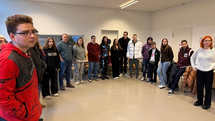 Eine Gruppe Studierender steht im Seminarraum im Halbkreis aufgestellt und schaut in die Kamera