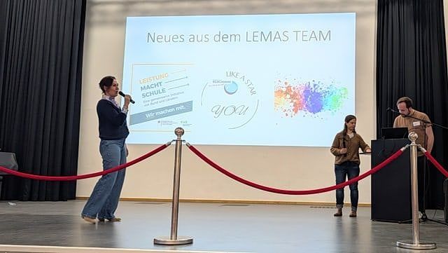 Drei Personen auf Schulbühne mit Bild von Lemas im Hintergrund auf Screen zu sehen