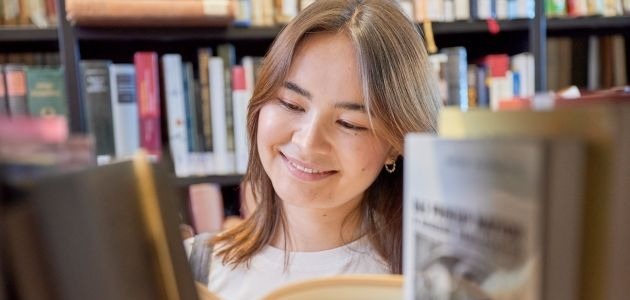Blick durch ein Regal auf eine lächelnde junge Frau, die auf ein Buch im Regal schaut