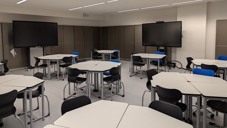 Zu erkennen ist der neu ausgestattete R05. Wir sehen Fünfektische in Form von Gruppentischen zusammengestellt. An den Seiten sind zwei Displays zu erkennen.