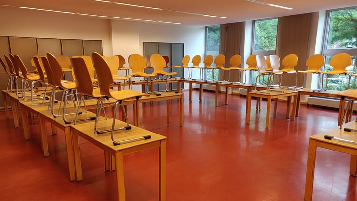 Zu erkennen ist ein klassischer Seminarraum. Der Boden ist Knallrot, die Standard-Tische in einem frontalen Setting in U-Form ausgerichtet. Es handelt sich um das "Vorher"-Bild aus R05.