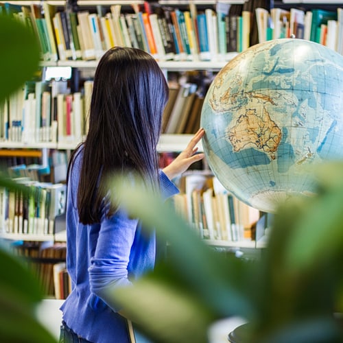 Eine Frau steht in einer Bibliothek mit dem Rücken zu der Kamera und zeigt etwas auf einem Globus.