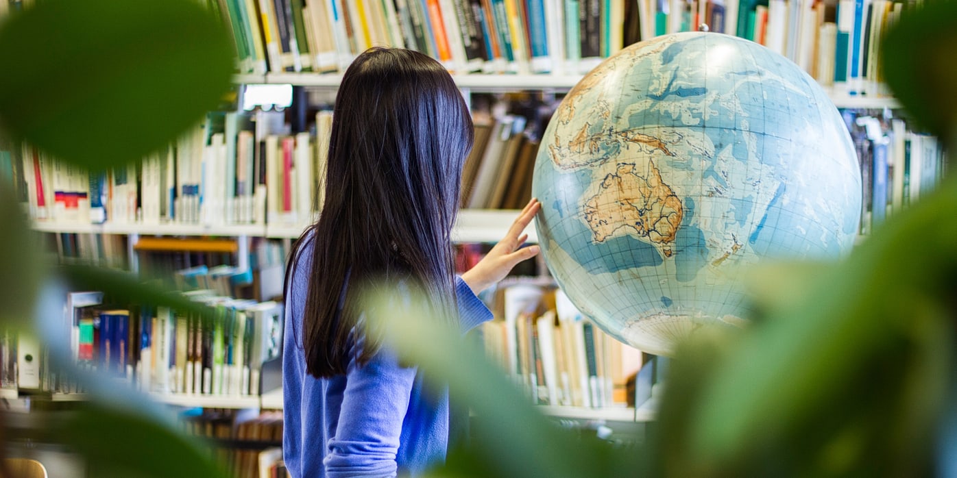 Eine Frau steht in einer Bibliothek mit dem Rücken zu der Kamera und zeigt etwas auf einem Globus.