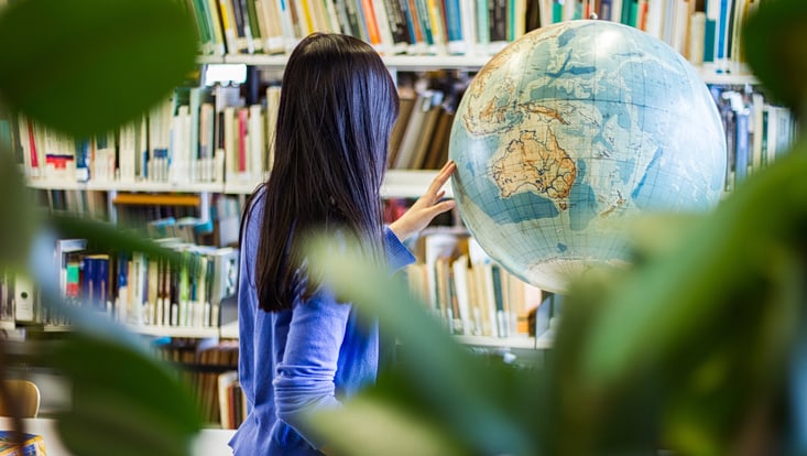 Eine Frau steht in einer Bibliothek mit dem Rücken zu der Kamera und zeigt etwas auf einem Globus.