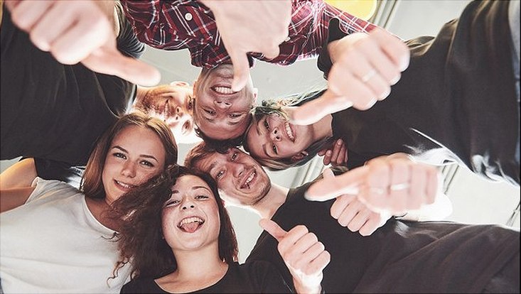 Eine Gruppe steht lachend im Kreis, den Blick nach unten in die Kamera gesenkt