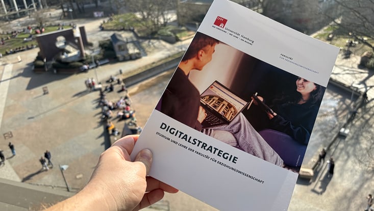 Zu sehen ist eine gedruckte Version der Digitalstrategie der Fakultät für Erziehungswissenschaft. Im Hintergrund ist ein belebter Campus zu erkennen.