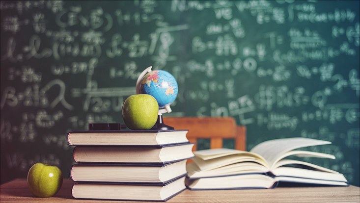 Bücherstapel auf Schreibtisch mit Mini-Globus und zwei grünen Äpfeln vor mit Formeln vollgeschriebener Tafel