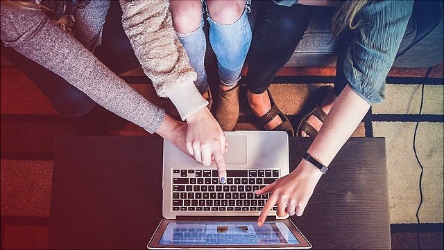 aufgeklappter Laptop von oben, drei Menschen davor, nur Arme und Beine zu sehen, gestikulieren und zeigen auf Bildschirm