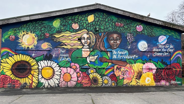 Eine mealte Hauswand in sehr bunten Farben. Das Bild zeigt einen großen Baum, darunter stehen eine weiße und eine schwarze Person, die gemeinsam eine Weltkugel halten. Das Bild zeigt verschiedene Schriftzüge auf Englisch, wie: "One Human Race"