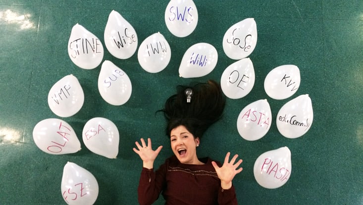 Eine junge Frau liegt am Boden zwischen weißen Luftballons, auf denen verscheidene unibezogene Abkürzungen wie z.B. VMP, SWS STiNE, WiSe stehen.