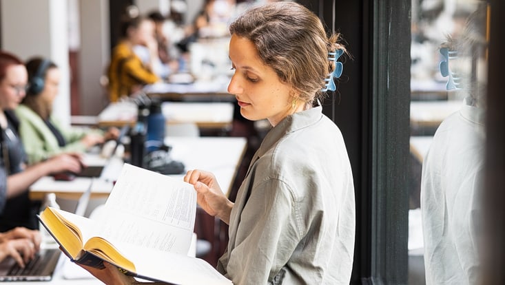 Studierende liest in der Bib am Fenster Eine Studierende sitzt in der Bibliothek auf der Fensterbank und liest ein Buch. Hinter ihr sind weitere Studierende zu sehen, die an Laptops arbeiten.