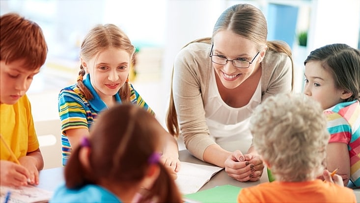 Eine lächelnde Lehrerin und Grundschulkinder sitzen um einen Tisch