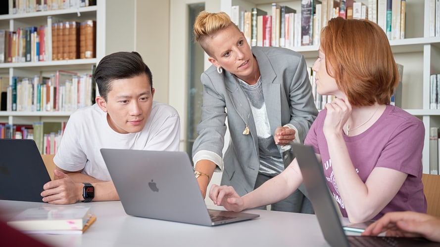 Lehrende mit zwei Studierenden vor Laptop Eine Lehrende steht zwischen zwei Studierenden, beugt sich über einen Laptop und erklärt etwas. Die Studierenden hören zu.