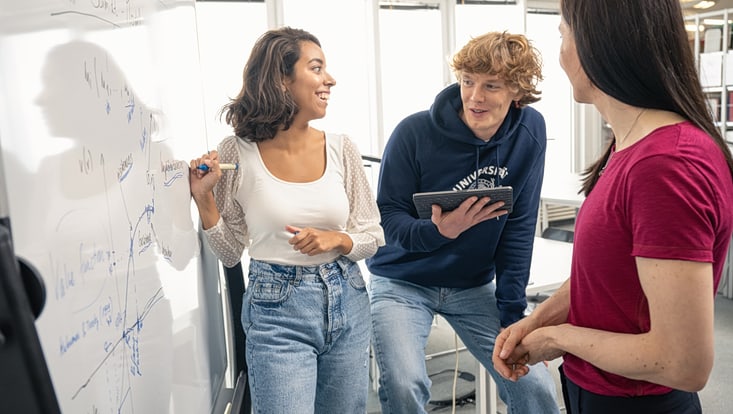 Eine Studierende schreibt etwas an ein Whiteboard, das ein Kommilitone mit einem Tablet ihr diktiert. Die Dozentin steht daneben und beobachtet die beiden.