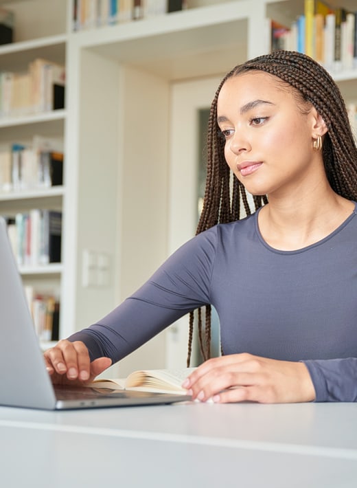 Eine junge Frau sitzt in einem Uniraum vor einem Laptop, hinter ihr sind Bücherregale zu sehen