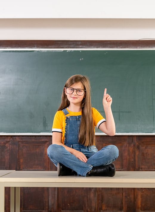 Ein kleines Mädchen sitzt auf einem Tisch vor einer Schultafel und meldet sich.