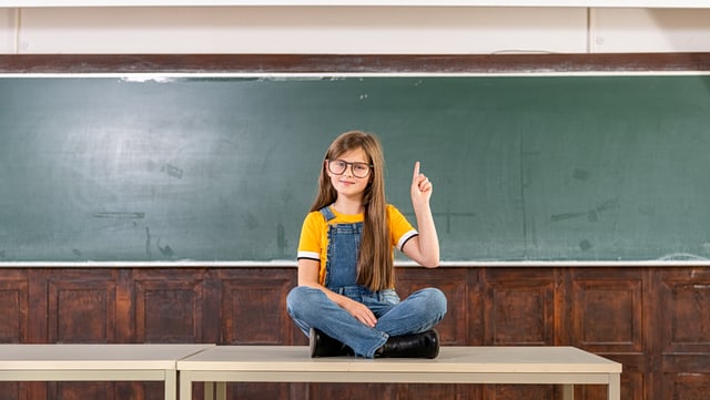 Ein kleines Mädchen sitzt auf einem Tisch vor einer Schultafel und meldet sich.
