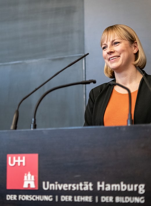 Eine Lehrende steht hinter einem Rednerpult, das das Logo der Universität Hamburg trägt, und spricht ins Mikrophon.