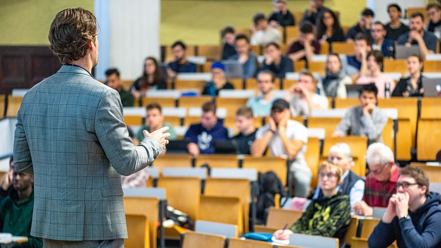 Ein Lehrender steht im Hörsaal und hält eine Vorlesung vor Studierenden.