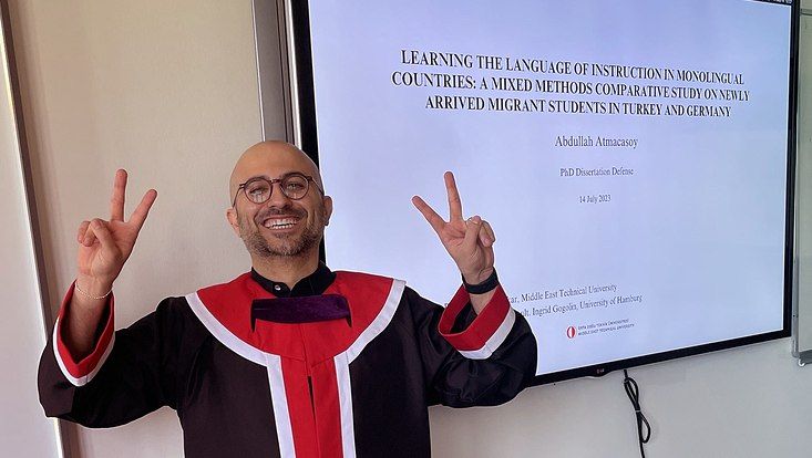 Abdullah Atmacasoy posiert im Doktor-Talar vor einem Smartboard mit dem Titel seiner Doktorarbeit.