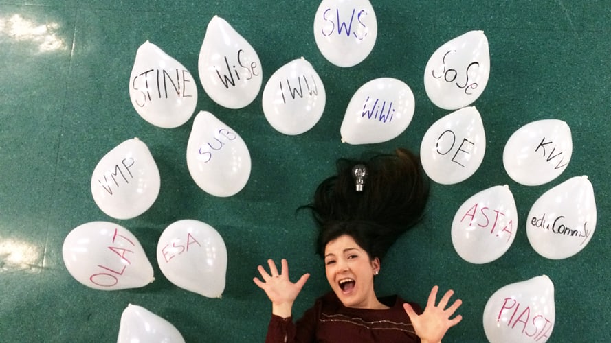 Eine junge Frau liegt am Boden zwischen weißen Luftballons, auf denen verscheidene unibezogene Abkürzungen wie z.B. VMP, SWS STiNE, WiSe stehen.