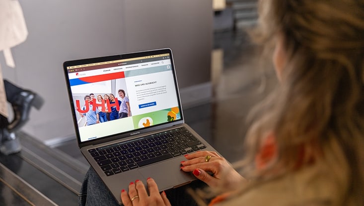 Eine Frau sitzt auf einer Treppe und hat einen Laptop auf dem Schoss. Man sieht sie nur von hinten, hat aber dafür den Bildschirm des Laptops mit einer geöffneten Webseite der Uni Hamburg im Mittelpunkt des Fotos im Blick..