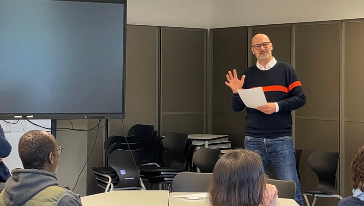 steckbrief-poeschke733x414 Dr. Jan Poerschke spricht vor den Alumni über seinen Werdegang. Dabei steht er in einem Seminarraum, in dem im Hintergrund ein ausgeschaltetes Smartbord erkennbar ist. Im vorderen Teil des Bildes erkennt man die Rückenansicht einiger Alumni.