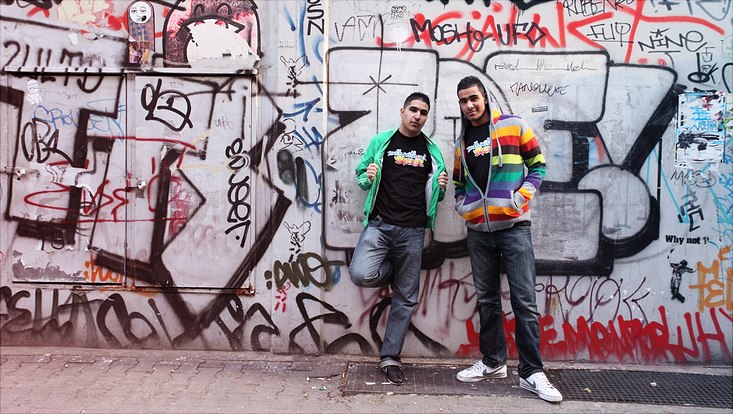 Zwei Jugendliche stehen vor einer mit Grafitti besprühten Mauer und schauen in die Kamera.