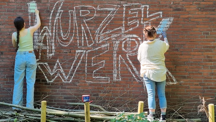20240513-122703-733x414 zwei Studierende gestalten mit Kreide den Schriftzug Wurzelwerk an einer Backsteinmauer