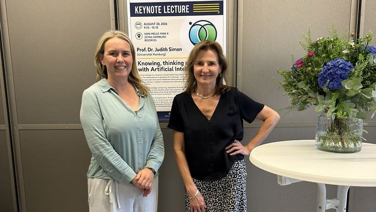 Prof. Dr. Judith Simon (links) und Prof Dr. Kerstin Michalik (rechts) stehen lächelnd vor einem Plakat, das Prof. Dr. Judith Simons Keynote Lecture "Knowing, Thinking and Being with Artificial Intelligence" bewirbt. Neben ihnen ein Strauß Blumen.