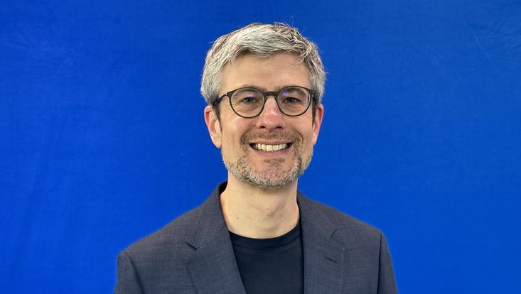 Portrait von Viktor Werner vor einer blauen Wand. Er trägt eine runde Brille, ein graues Jarkett und ein schwarzes T-Shirt. Er lächelt in die Kamera.