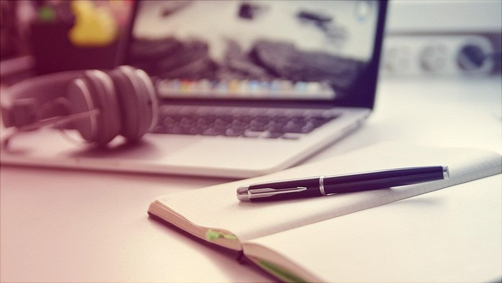 im Hintergrund ist ein Laptop zu sehen, im Vordergrund ein Notizbuch mit Füller