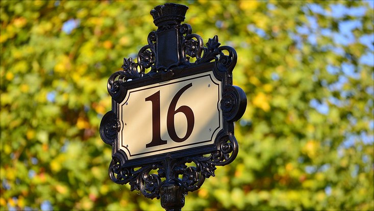 Auf einem verschnörkelten Straßenschild steht die Zahl 16, im Hintergrund sieht man einen verschwommenen Baum