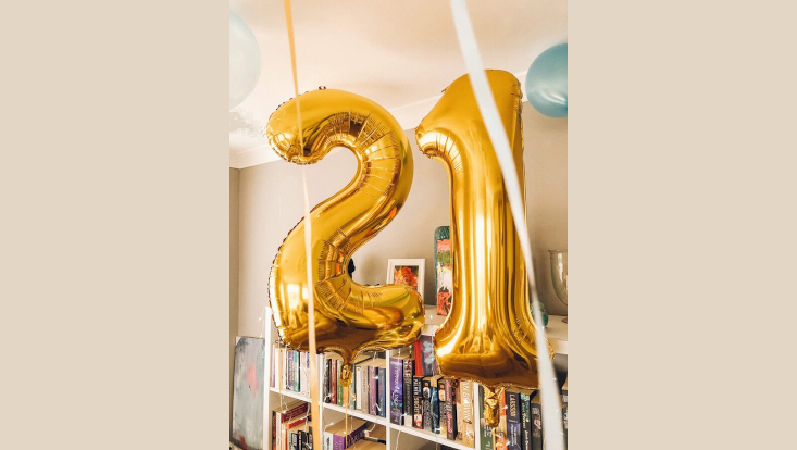 Zwei Heliumballons stellen die Zahl 21 dar. Im Hintergund steht ein Bücherregal und weitere Luftballons